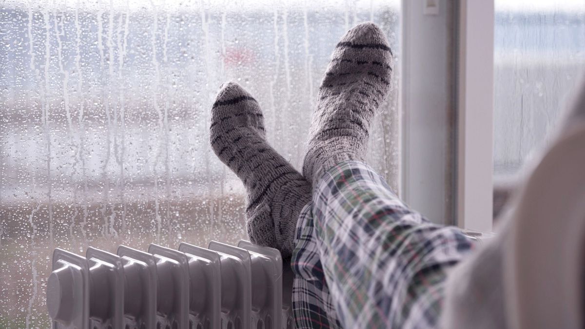 Female's feet with gray woolen socks heating on the oil radiator next to a home window in rainy weather.GettyImages