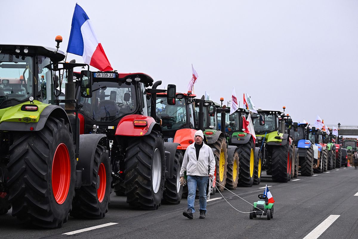 A farmer walks past lined-up tractors, holding a stuffed beet and pulling a toy tractor waving a French flag, as farmers coming from Meuse and Moselle block the A4 highway, near Jossigny, east of Paris on January 30, 2024 amid nationwide protests called by several farmers unions on pay, tax and regulations. Farmers protests across Europe are growing as they demand better conditions to grow produce and maintain a proper income. (Photo by Bertrand GUAY / AFP)