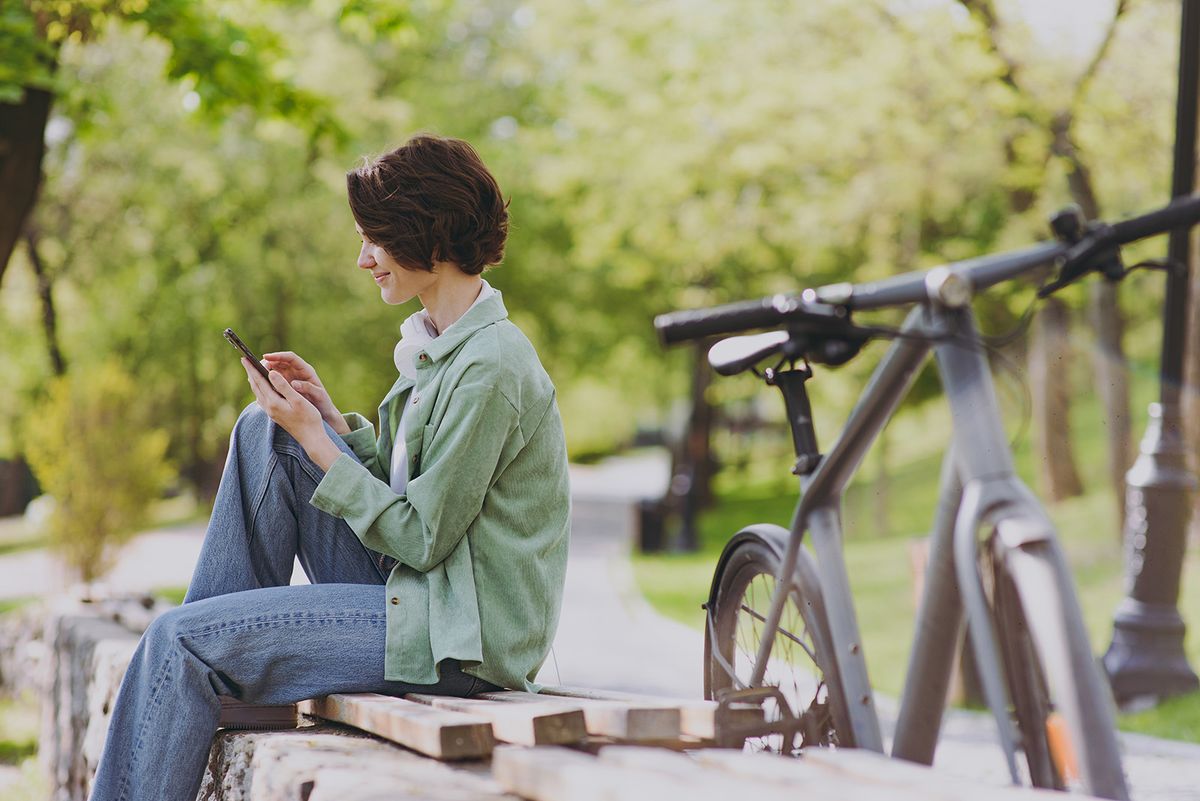 SIde view happy young woman 20s in green jacket jeans sit on bench near bicycle in city spring park outdoors hold mobile cell phone chat online in social network People active urban youthful concept.