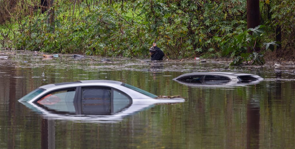 A man swims chest-deep through flood waters with his cell phone near three cars that are submerged after rain flooded several areas of Long Beach from the first in a series of storms hits Southern California