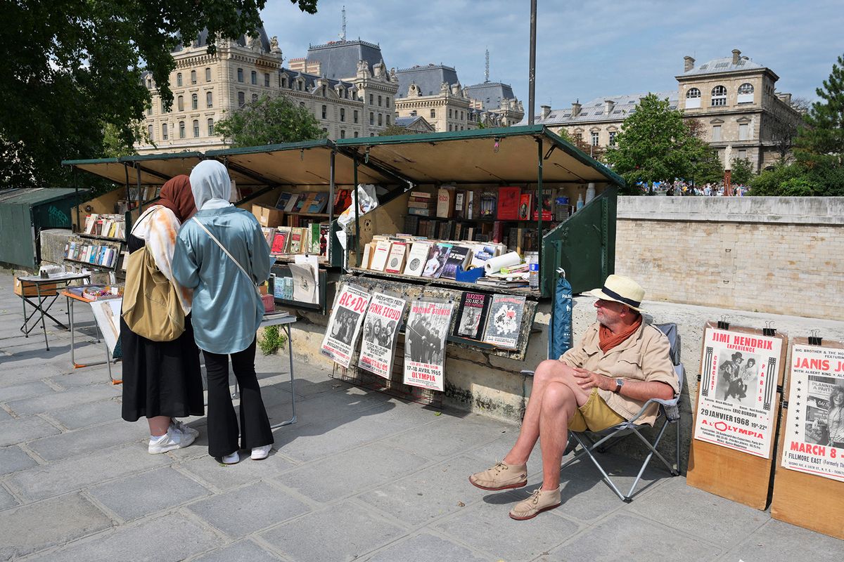 (FILES) Philippe Bayle (R), a 65-year-old bouquinist (bookseller sits at his stand, selling antiquarian books and old posters, on the left bank of the Seine River in Paris on August 29, 2023. French President Emmanuel Macron has decided to abandon the planned movement of the Parisian booksellers' boxes installed on the banks of the Seine in preparation for the opening ceremony of next summer's Olympic Games in Paris, the Elysee announced on February 13, 2024. (Photo by MIGUEL MEDINA / AFP)