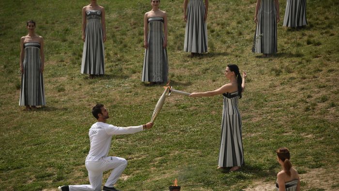 Az első fáklyahordozó, a 2020-as olimpiai aranyérmes evezős Stefanos Ntouskos kapja a lángot a főpapnőt alakító Mary Mina görög színésznőtől