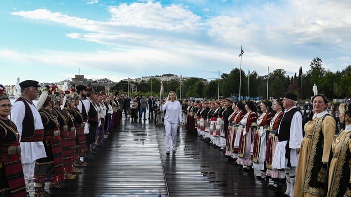 A görög olimpiai diszkoszvető, Tasoula Kelesidou áthalad a hagyományos görög viseletet viselő résztvevők között.