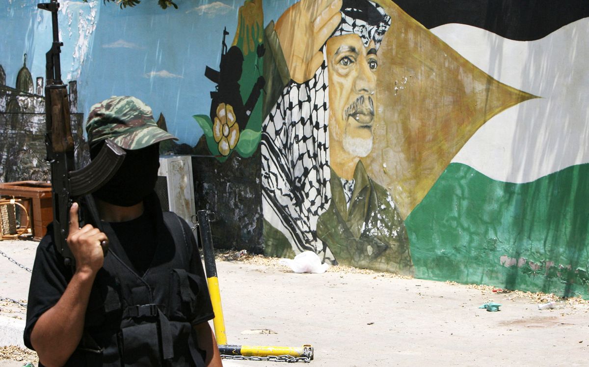 A masked Hamas gunman stands guard outside the presidential compound bearing a portrait of late Palestinian leader Yasser Arafat in Gaza City, 16 June 2007. Hamas cemented its control over Gaza today, seizing weapons from the routed security services of their Fatah rivals, as the home of iconic leader Arafat became the latest target of looting in the territory. For a second day in a row, Hamas fighters raided the homes of security personnel across the territory, confiscating weapons as they consolidated their victory after a week of bloody street battles, witnesses said. AFP PHOTO/MAHMUD HAMS (Photo by MAHMUD HAMS / AFP)