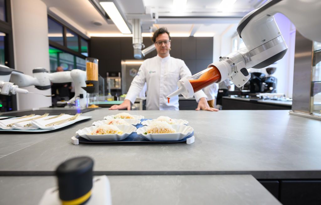 15 April 2024, Saxony, Dresden: Chef Benjamin Biedlingmaier, looks at a collaborative robot preparing a salad at the opening of the robot kitchen "Cetibar" in the CeTI Cluster of Excellence in the Barkhausen Building at Dresden University of Technology. In future, humans should be able to interact with robots or machines in real time. Research into this is being carried out in the special laboratory at the Technical University of Dresden. Photo: Robert Michael/dpa (Photo by Robert Michael/picture alliance via Getty Images)