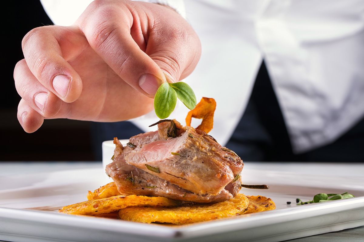 Chef in hotel or restaurant kitchen cooking, only hands. Prepared meat steak with potato or celery pancakes.He is working on the herb decoration.