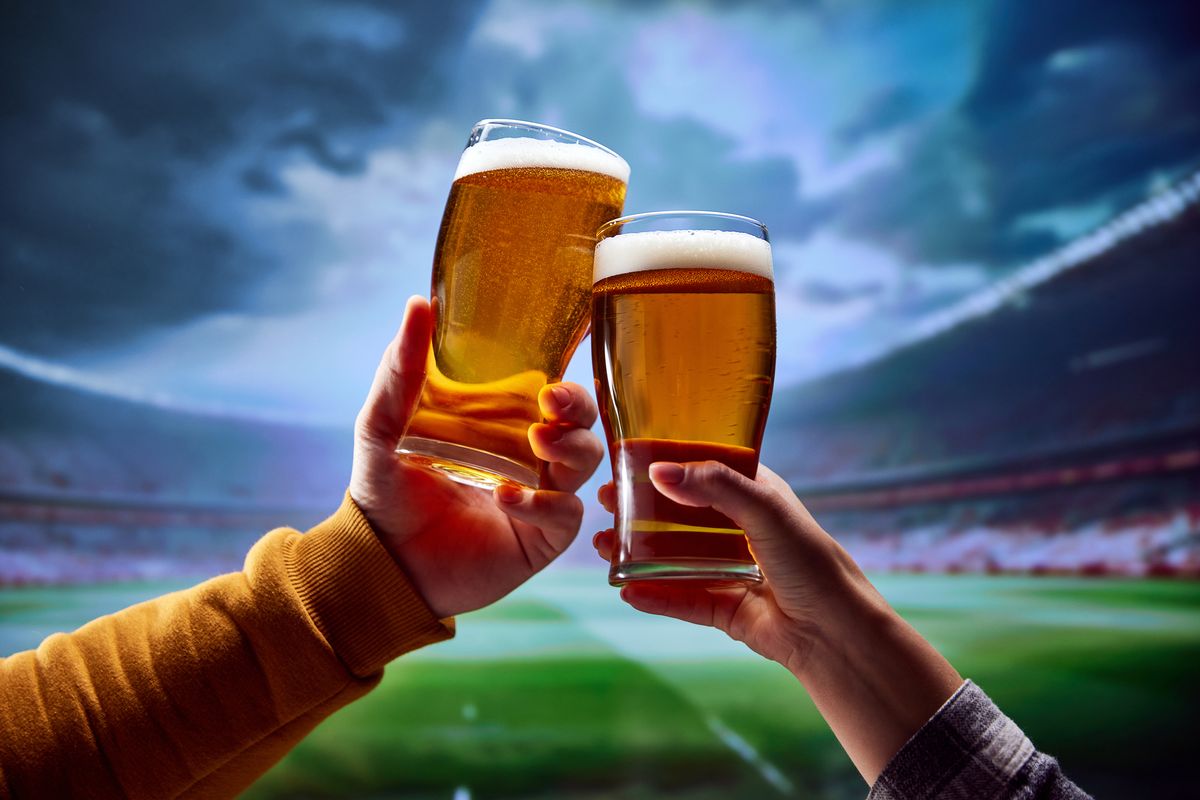 Two friends sitting at arena and drinking cold refreshing foamy beer and support their favorite football team during deciding match.