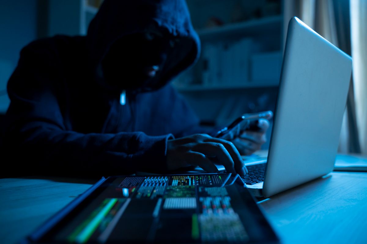 Anonymous person in the hood sitting in front of computer working with laptop and mobile phone