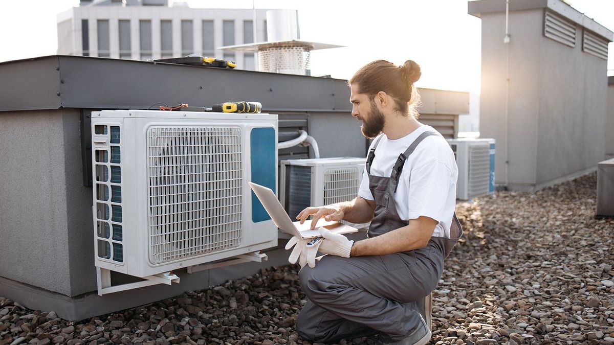 Side view of professional repairman wearing overalls and using modern laptop while looking at structure of air conditioners on factory roof. Concept of people, gadgets and maintenance.