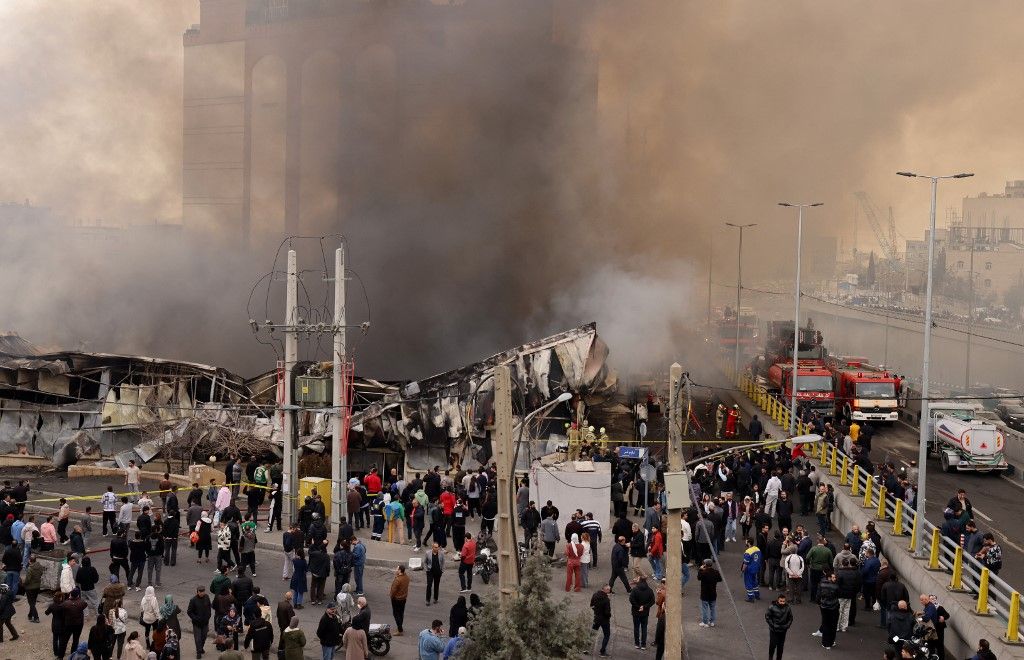 People stand and watch as firefighters battle a fire that broke out in Jannat Bazaar, west of Tehran on February 3, 2026. A massive fire broke out on February 3 at a bazaar west of the Iranian capital, state media reported, although the cause of the blaze