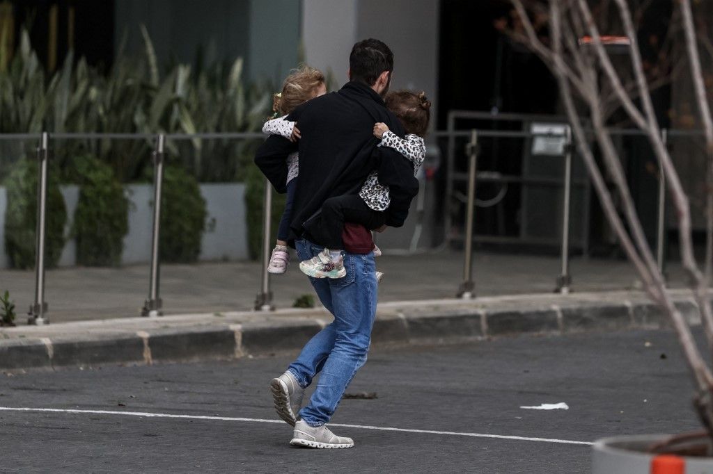 A man carries two children as he runs to take shelter after sirens sounded in Tel Aviv on February 28, 2026, following the announcement that Israel had launched a “preemptive strike” on Iran. President Donald Trump urged Iranians on February 28 to take ov
